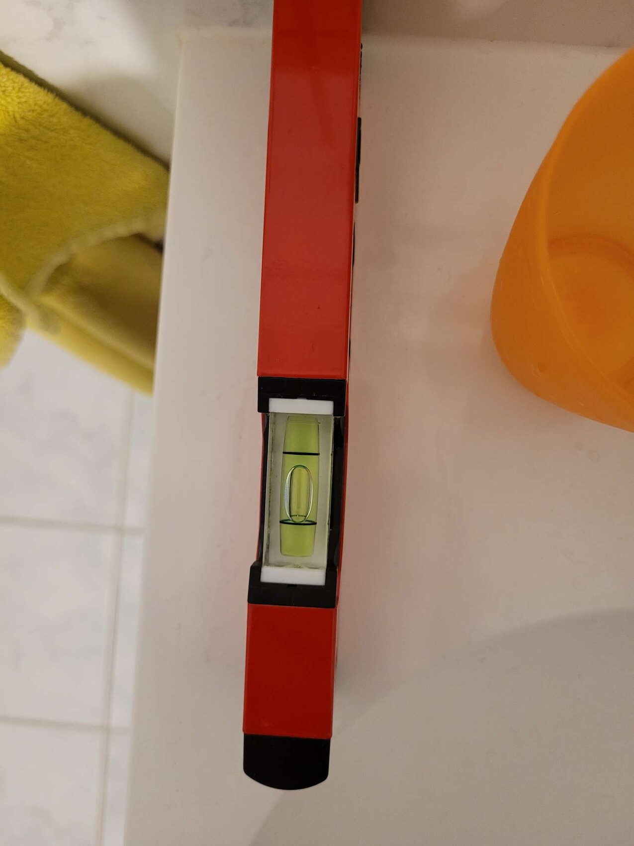Rote Wasserwaage auf weißer Badoberfläche neben gelbem Handtuch und orangem Becher.