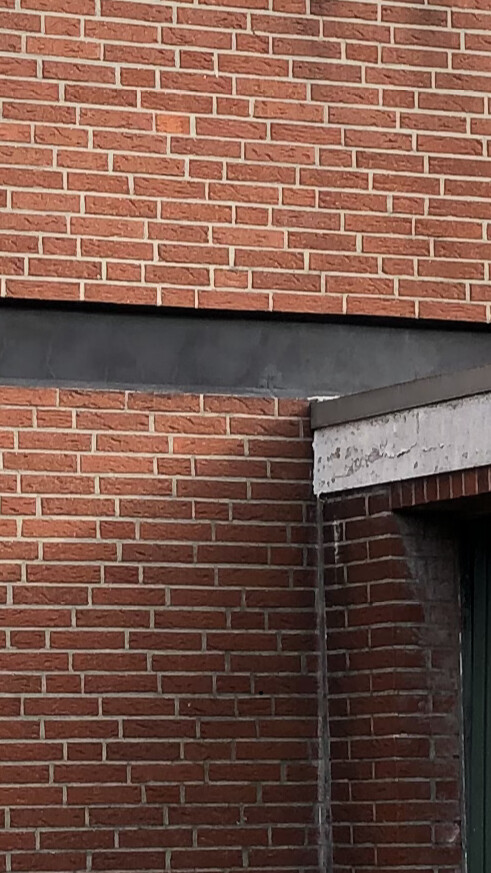 Außenansicht einer roten Ziegelwand mit schwarzem Querriegel; rechts Türrahmen mit grünem Türblatt.