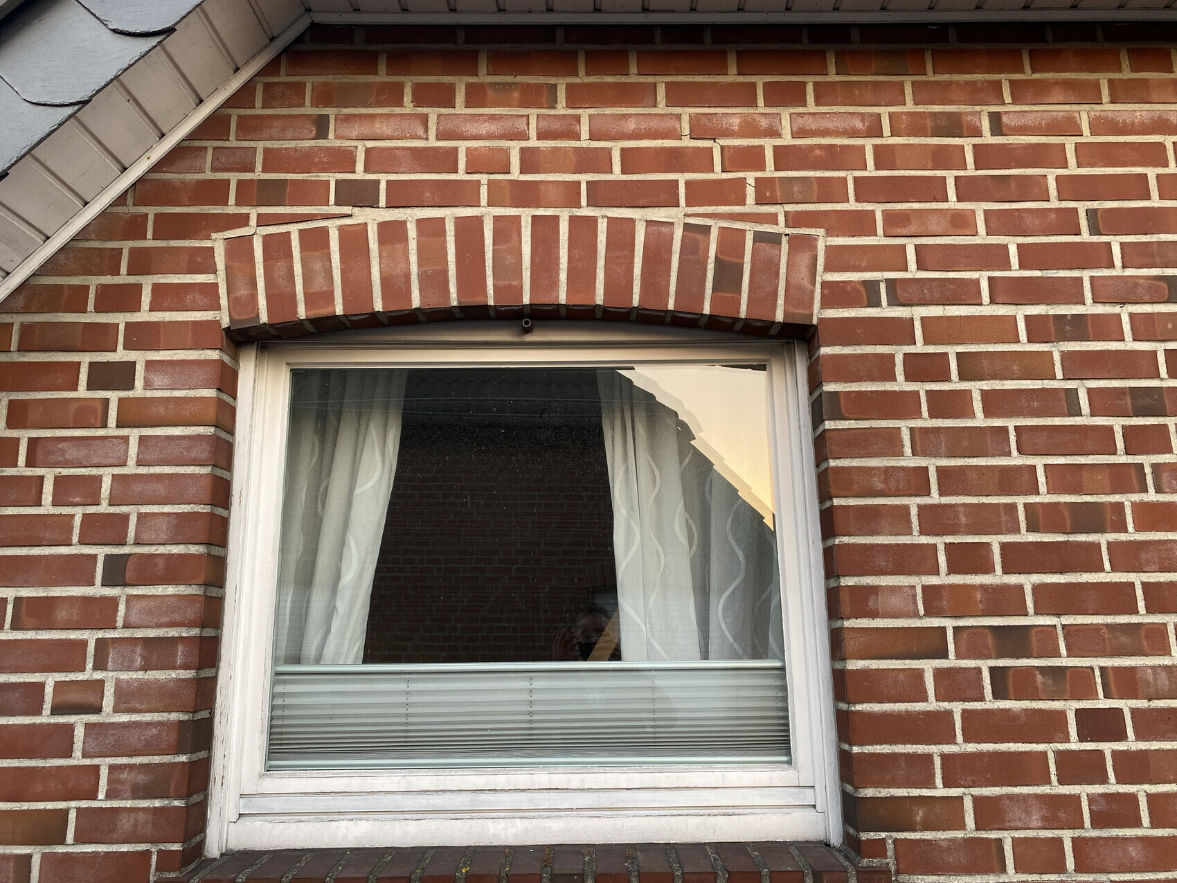 Fenster in roter Backsteinwand, weiße Vorhänge, horizontale Jalousien unten, Backsteinbogen darüber.