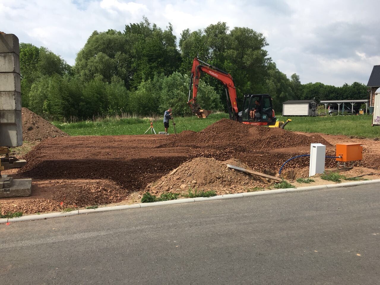 orangener Bagger gräbt Erde auf einer Baustelle, Haufen Erde, grüne Bäume im Hintergrund
