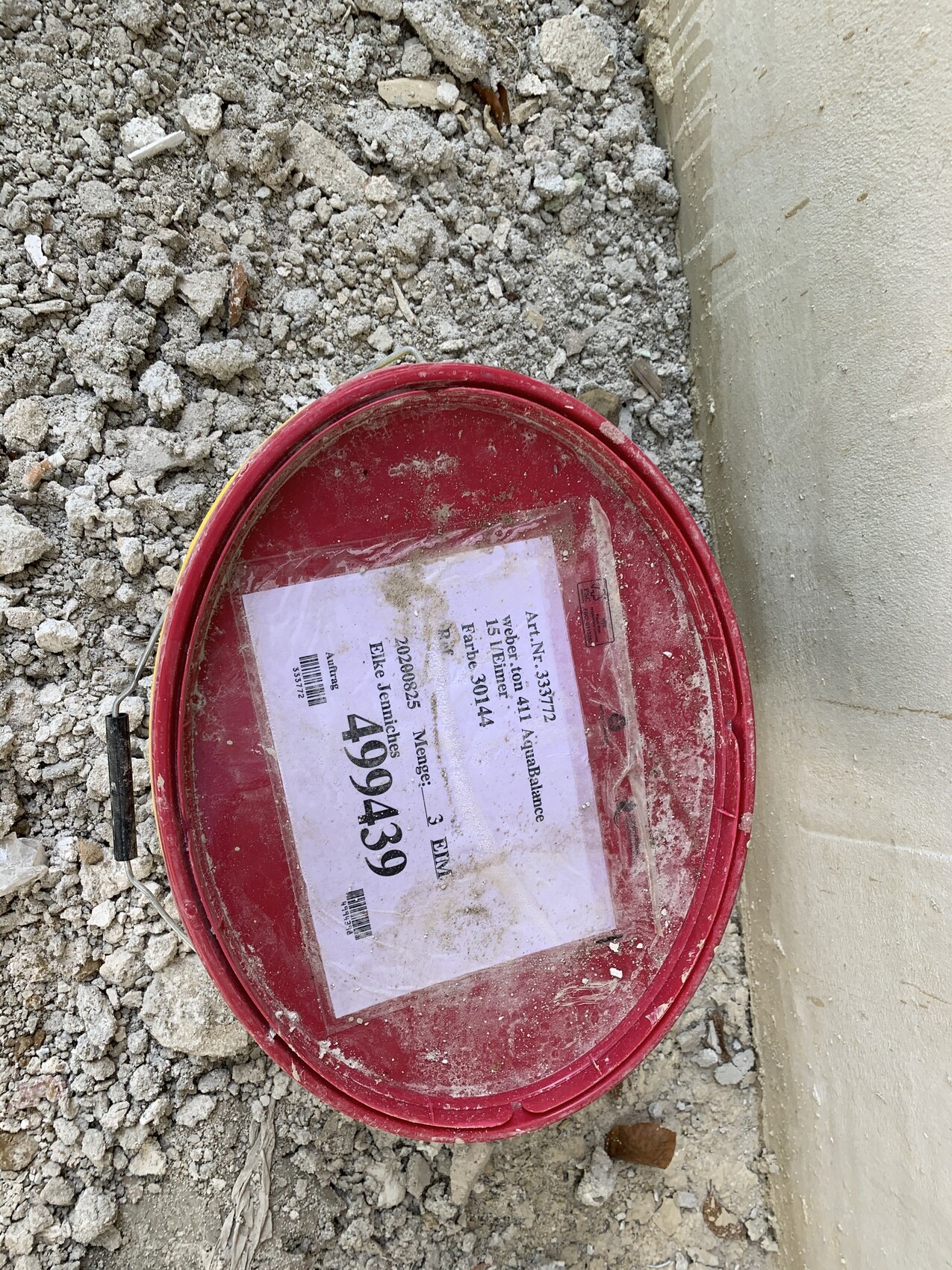 Roter Eimer mit beschriftetem Zettel liegt auf Bauschutt neben einer Wand.