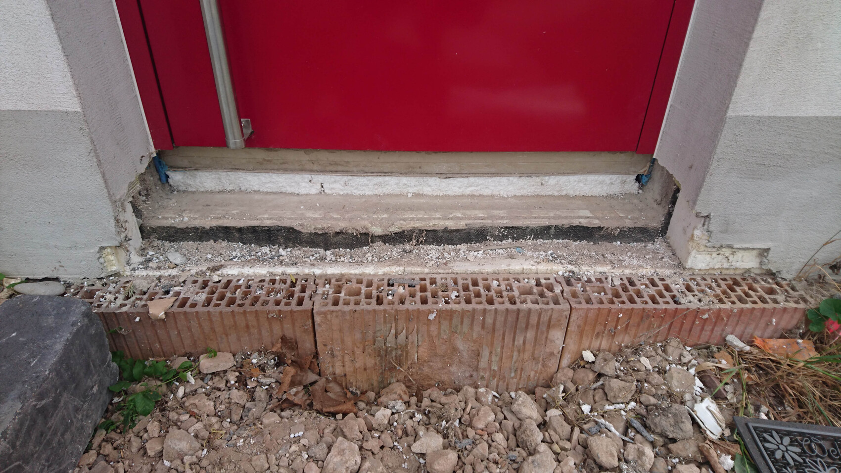 Rote Haustür am Eingangsbereich mit Baugrund, Bruchsteinmauer und Schutt davor.