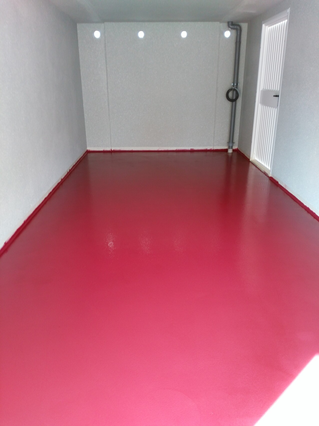 Kleiner, leerer Raum mit pinkem Boden, grauen Wänden und vertikaler Rohrleitung mit Ringgriff rechts.
