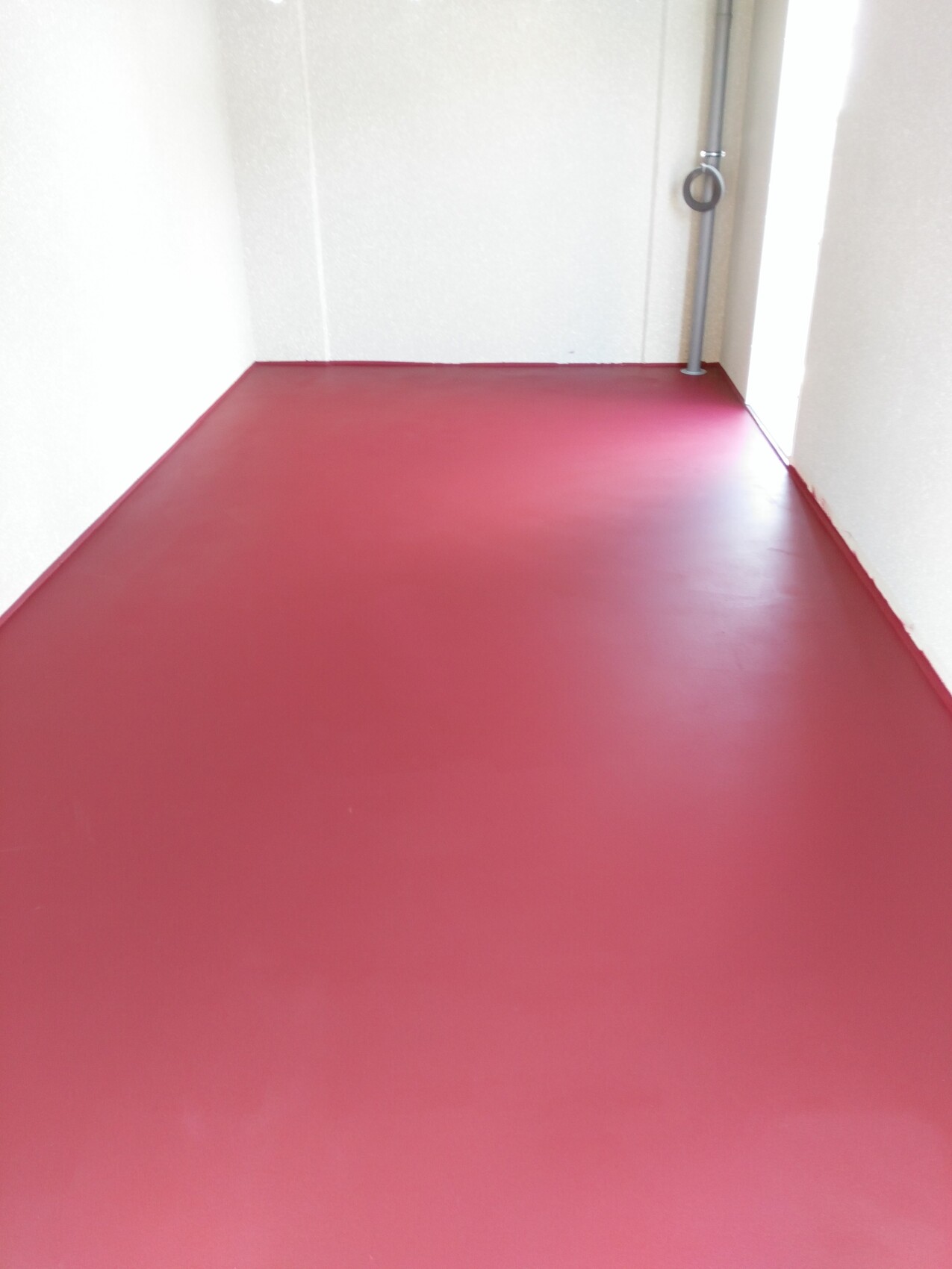 Korridor mit rotem, glänzendem Boden und weißen Wänden; ein Metallrohr mit Ring an der rechten Wand.