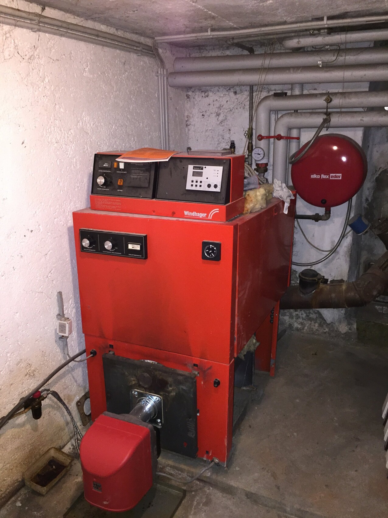 Roter Heizkessel im Kellerraum mit Rohren, Warmwassersystem und Messgerät.
