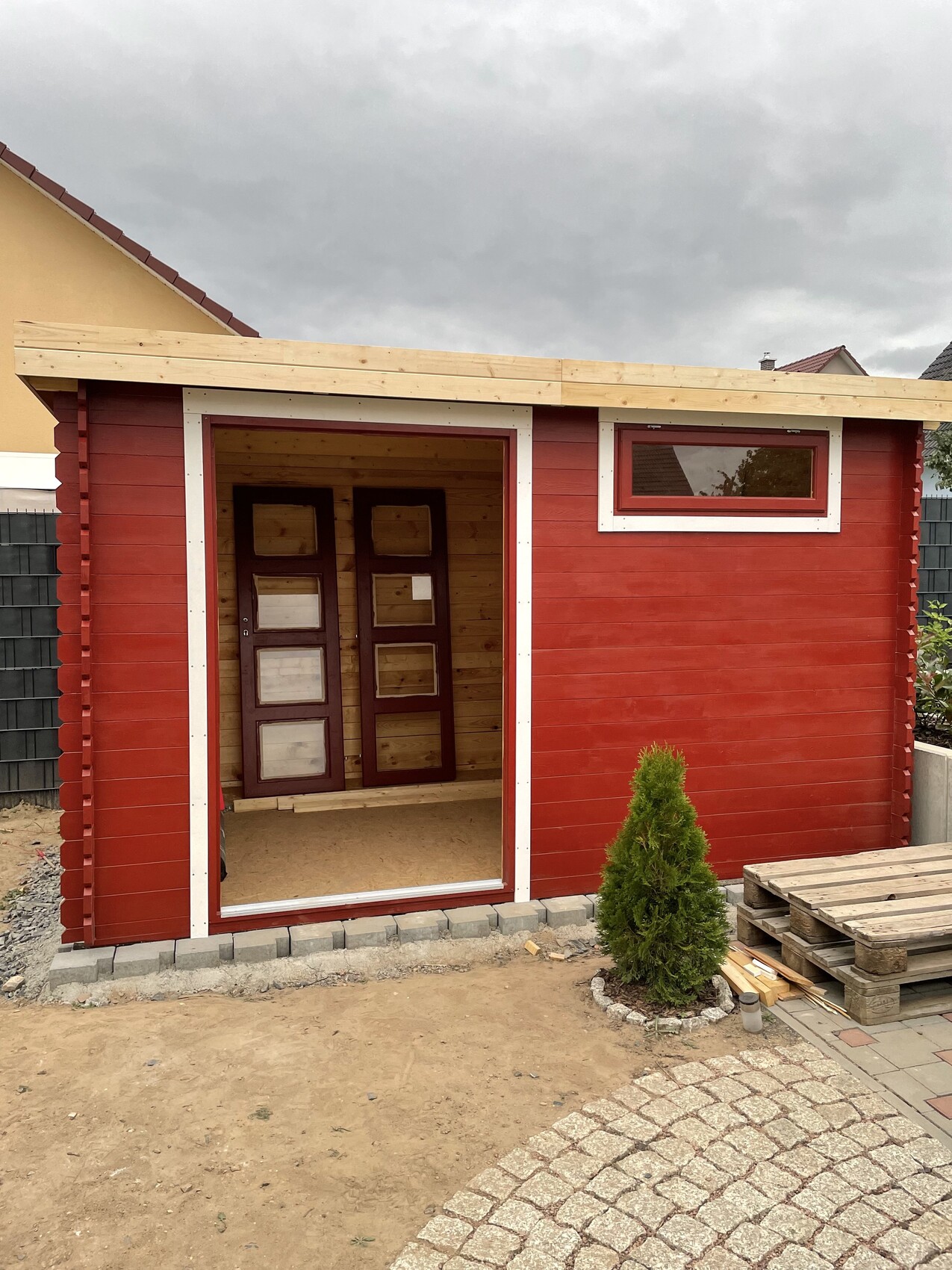 Kleiner roter Holzschuppen mit offener Tür im Garten, Paletten daneben.