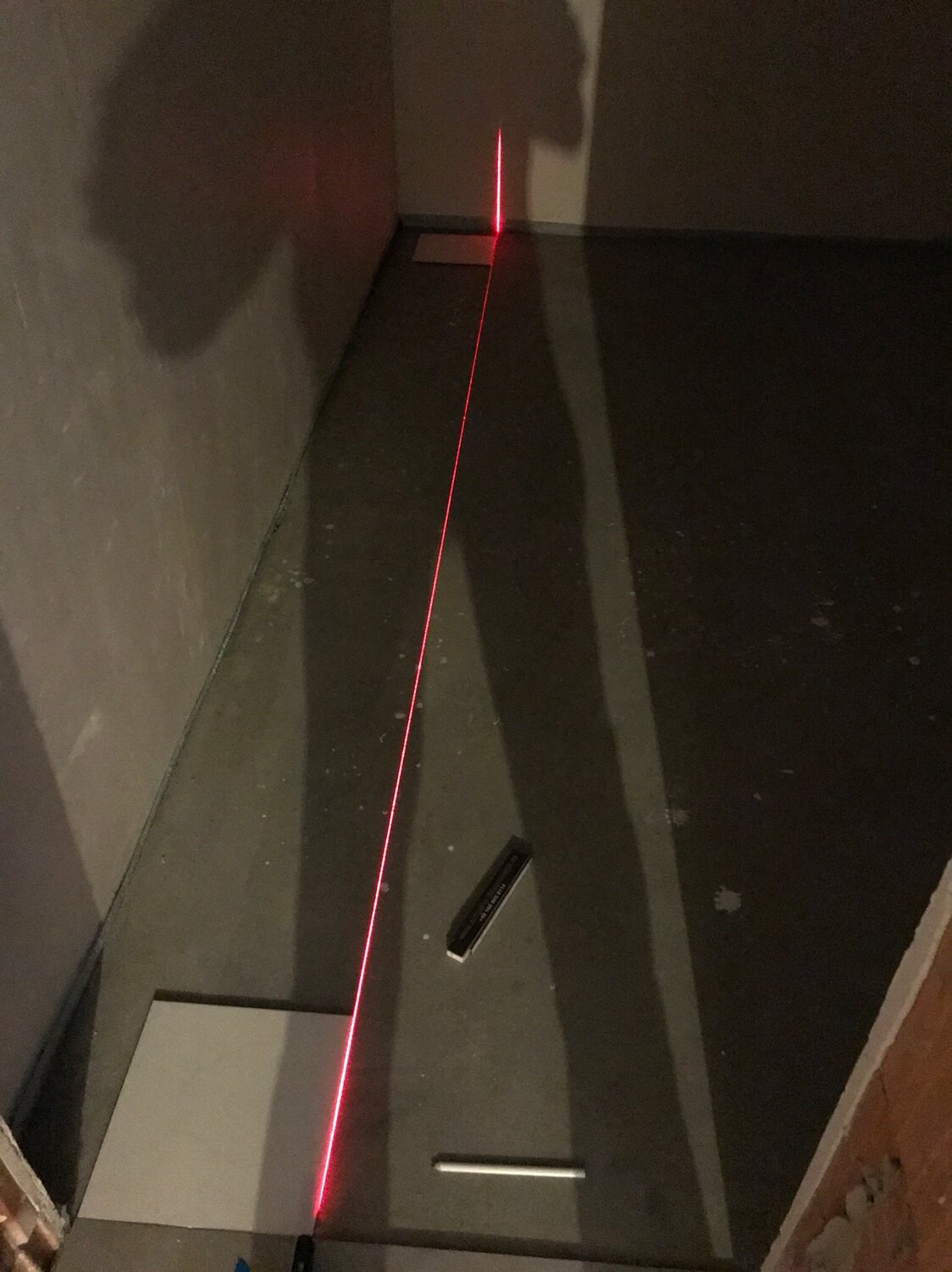 Diagonal roter Laserstrahl verläuft über dunklen Boden, Quelle ist ein Laser-Nivelliergerät am Boden.