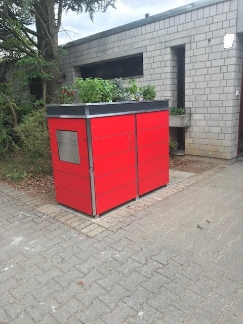 Roter Müllcontainer aus Metall vor grauem Gebäude auf einem gepflasterten Vorplatz.