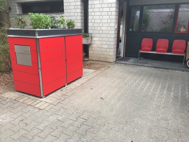 Roter Müllcontainer im Vorhof neben Eingangsbereich und roten Sitzbänken.
