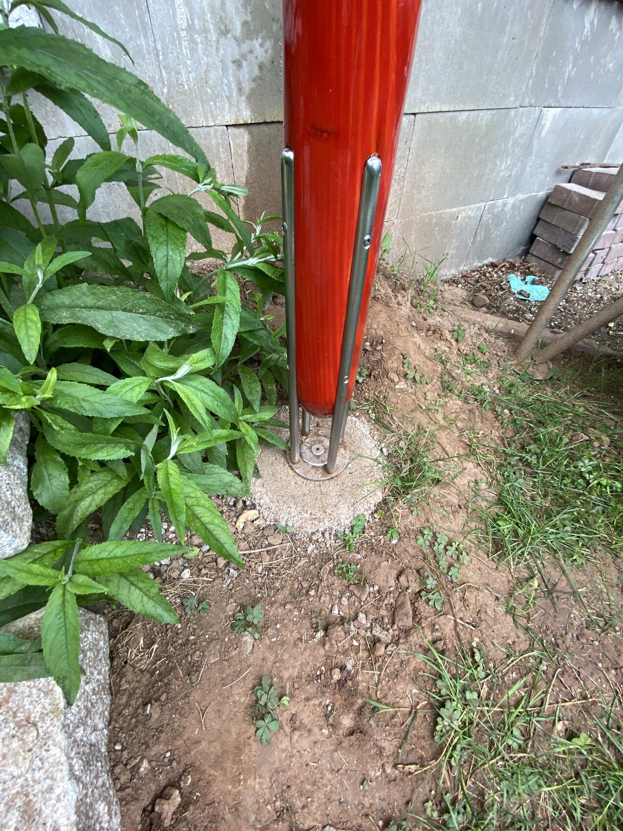 Roter vertikaler Pfosten mit Metallhalterungen auf runder Betonbasis, umgeben von Erde und Pflanzen.