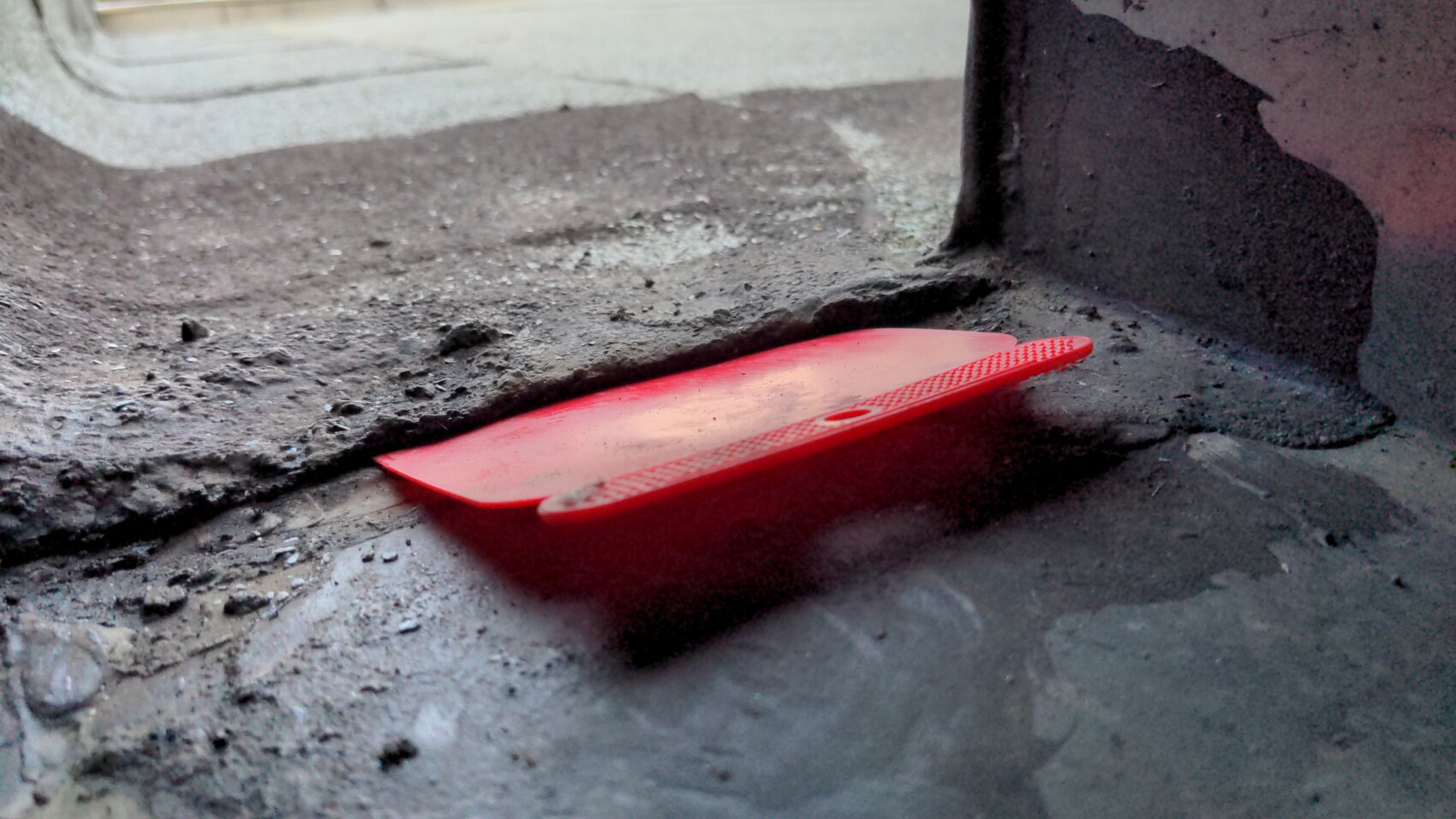 Roter Kunststoffspachtel liegt auf staubigem Betonboden neben einer dunklen Stütze.
