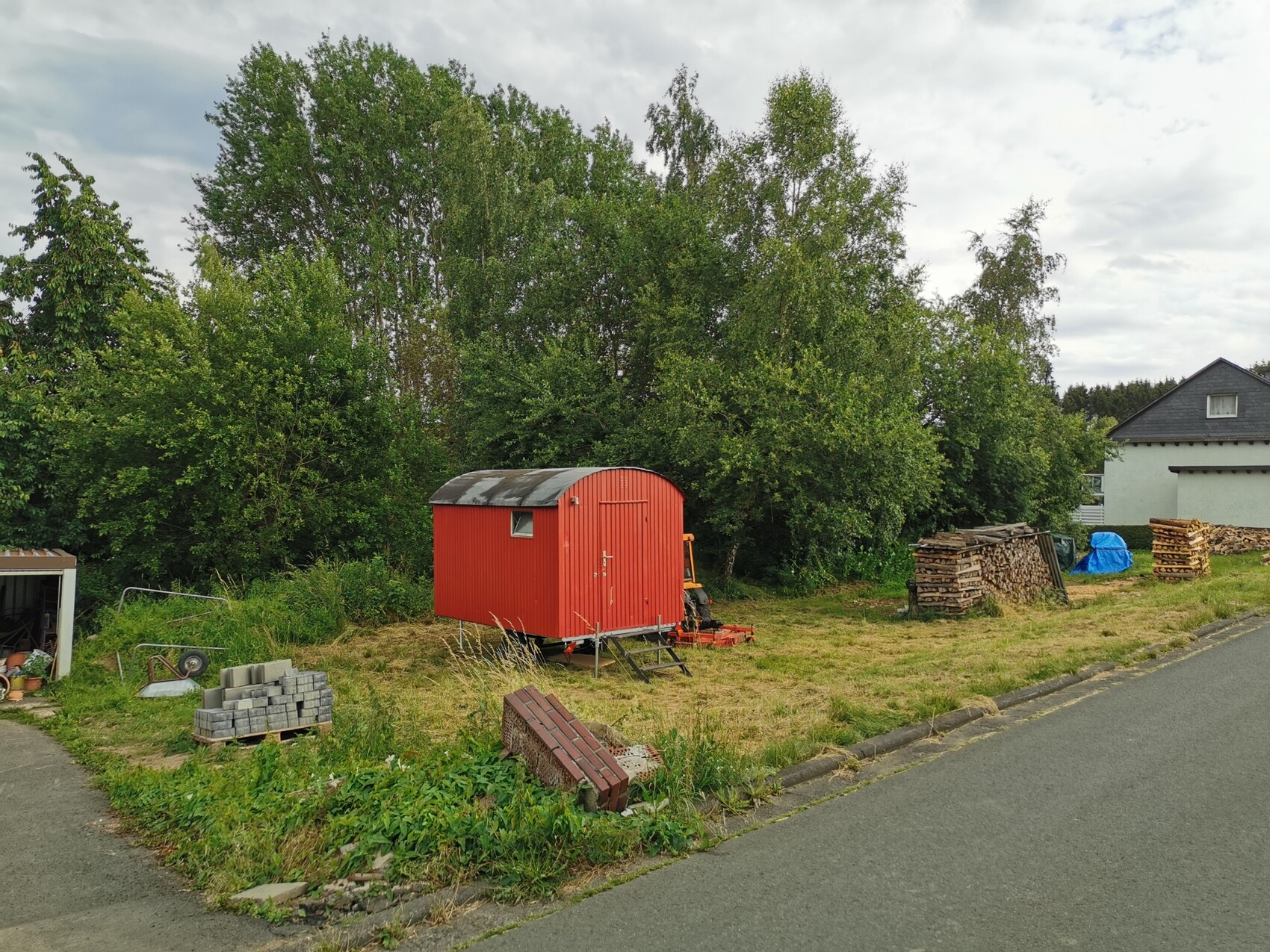 Roter Garten-Schuppen mit gewölbtem Dach, daneben Holzstapel und Bäume im Hintergrund