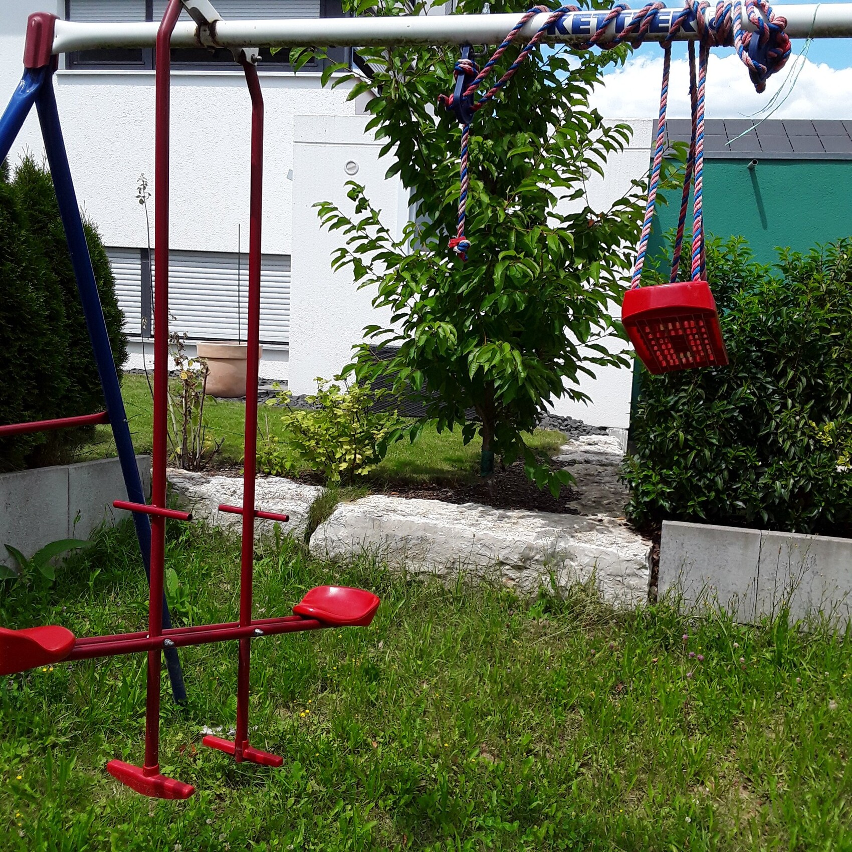 Rotes Schaukelgestell im Garten, rote Sitze hängen an Seilen; Baum, Hecke und Haus im Hintergrund.