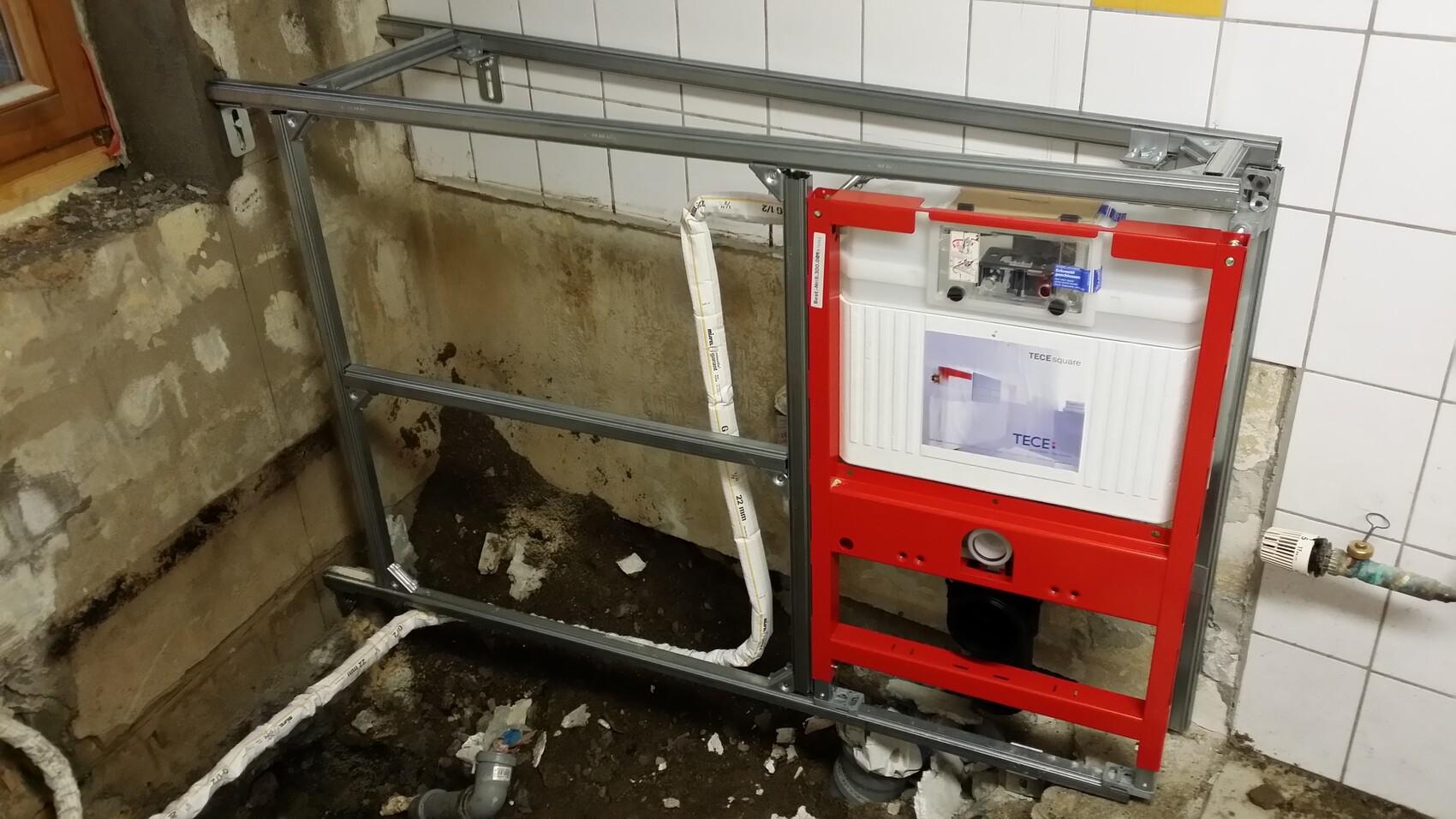 Wandhängendes WC-Traggestell aus Metallrahmen mit rotem Spülkasten vor Fliesenwand im Bad.