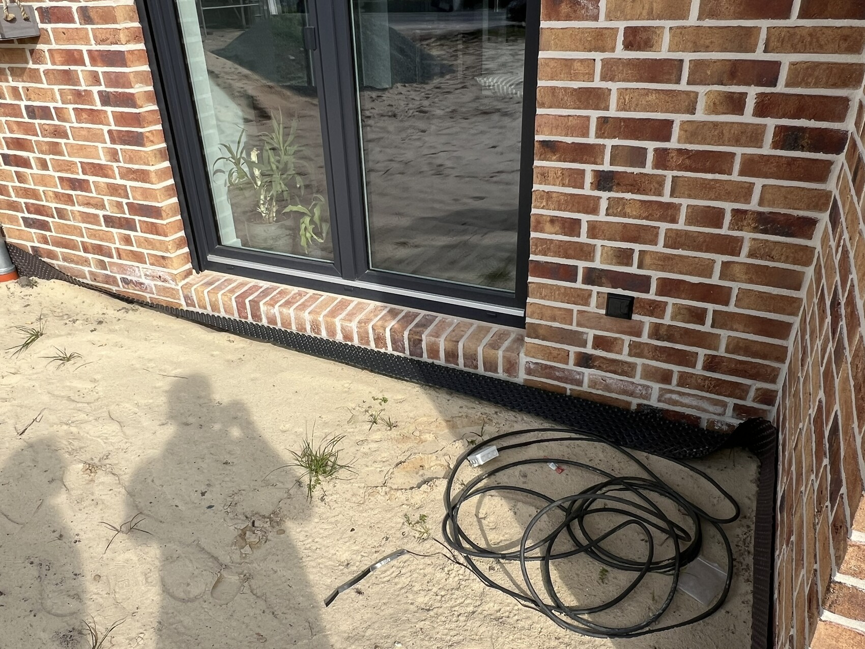Außenwand aus roten Ziegeln mit Glas-Schiebetür; Kabel und Schatten auf sandigem Boden.