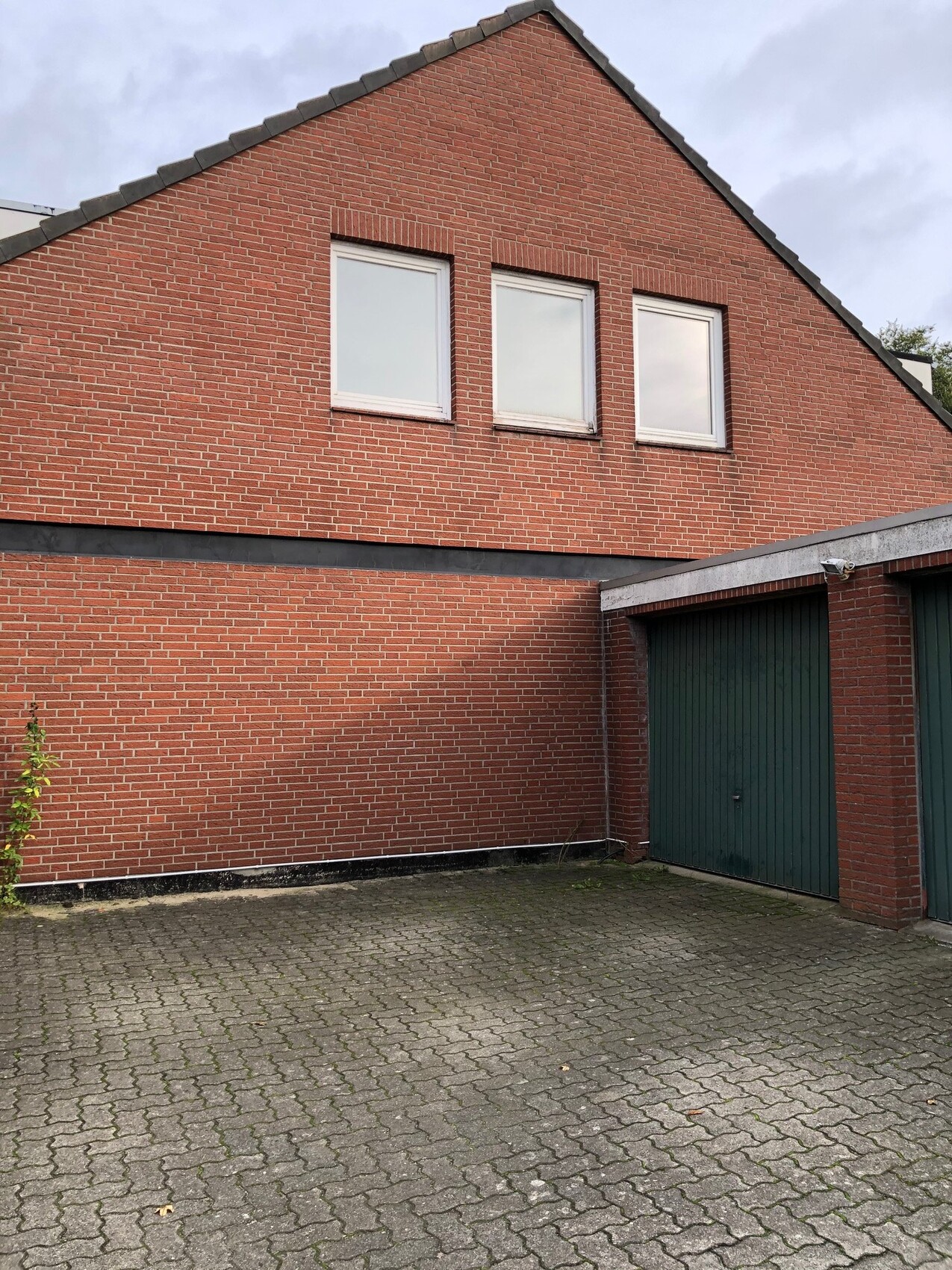 Ziegelhaus mit Giebel, drei Fenster im Obergeschoss, grüne Garagentore, gepflasterter Hof.