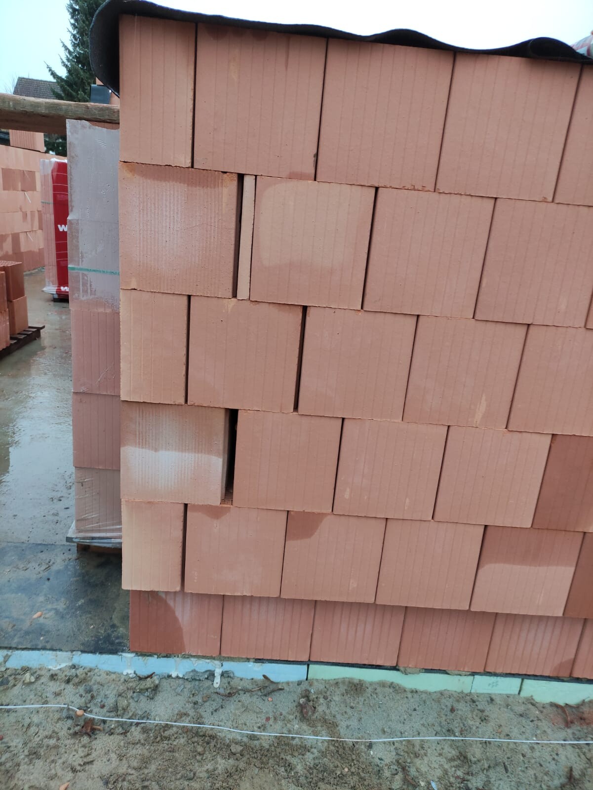 Stapel roter Backsteine auf Paletten an einer Baustelle, nasser Boden sichtbar.