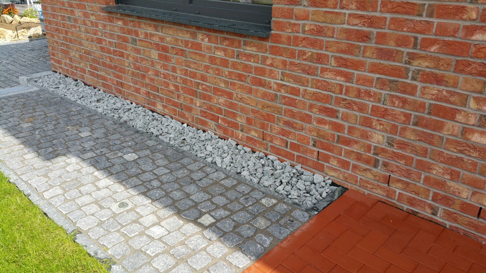 Außenansicht eines roten Backsteingebäudes mit Kiesstreifen am Fundament und gepflastertem Weg.