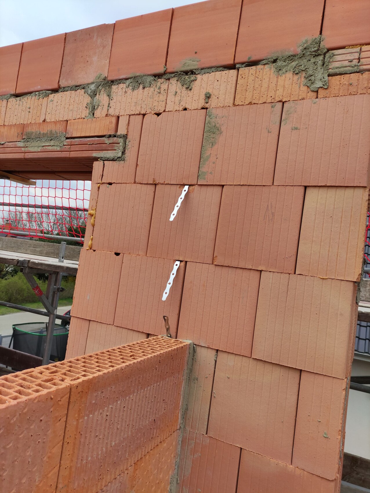 Baustelle: Wand aus roten Tonziegeln wird gebaut, Mörtelreste oben, Gerüstnetz im Hintergrund.