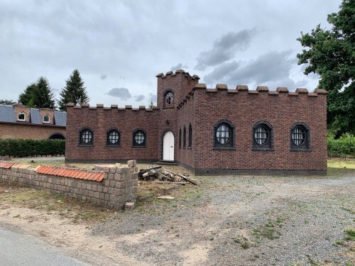 Kastellartiges rotes Backsteingebäude mit Zinnen, Rundturm, weiße Tür und Bogenfenstern, Kiesfläche.