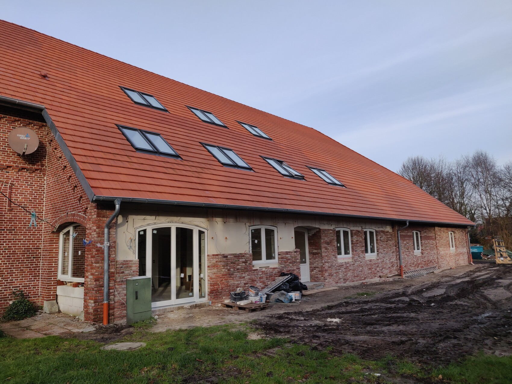 Langgestrecktes Haus mit rotem Ziegeldach, Dachfenstern und Baustellenmaterial davor.