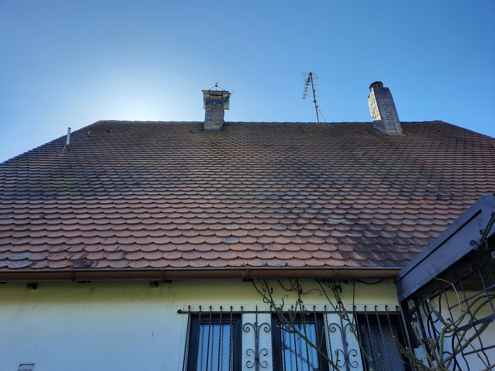 Dach mit roten Ziegeln, zwei Schornsteine, Antenne, blauer Himmel, Fenster mit Gitter.