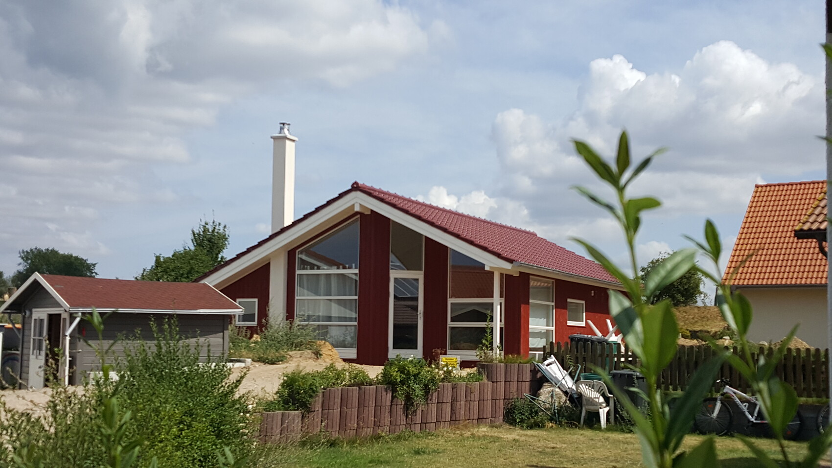 Rotes Einfamilienhaus von außen mit Garten und Nebengebäuden vor blauem Himmel.