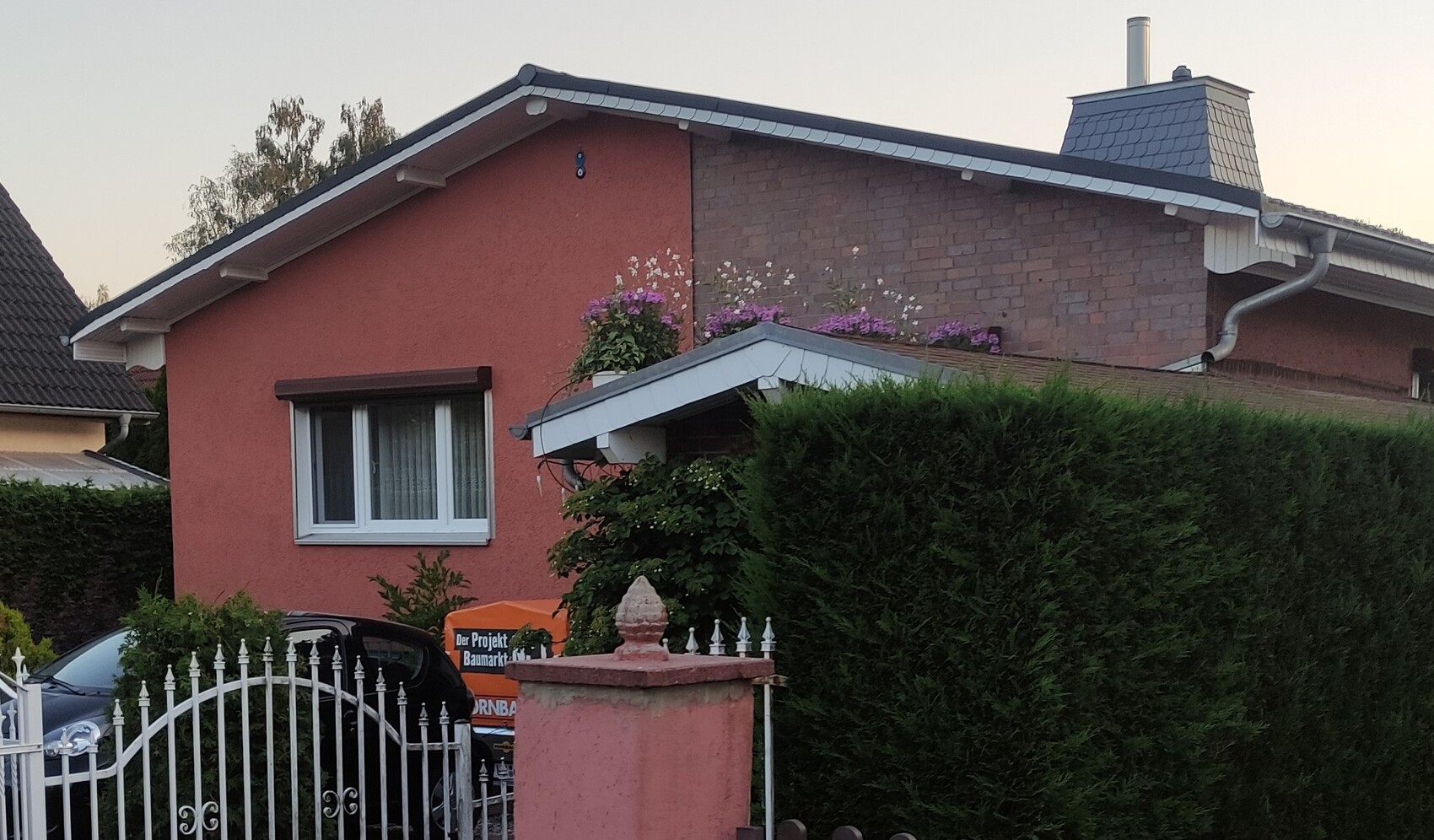 Rotes Einfamilienhaus mit weißen Fensterrahmen, Blumen am Vordach und dichte Hecke.