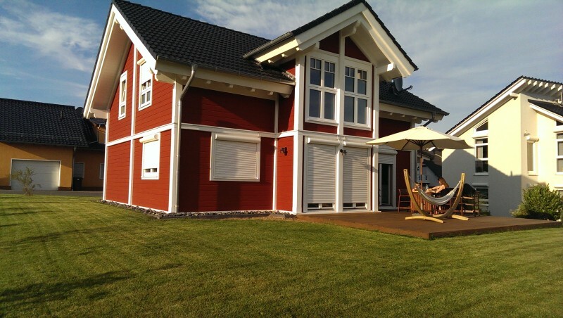 Rotes Holzhaus mit weißer Verzierung, Terrasse, Sonnenschirm und Hängematte im Garten.