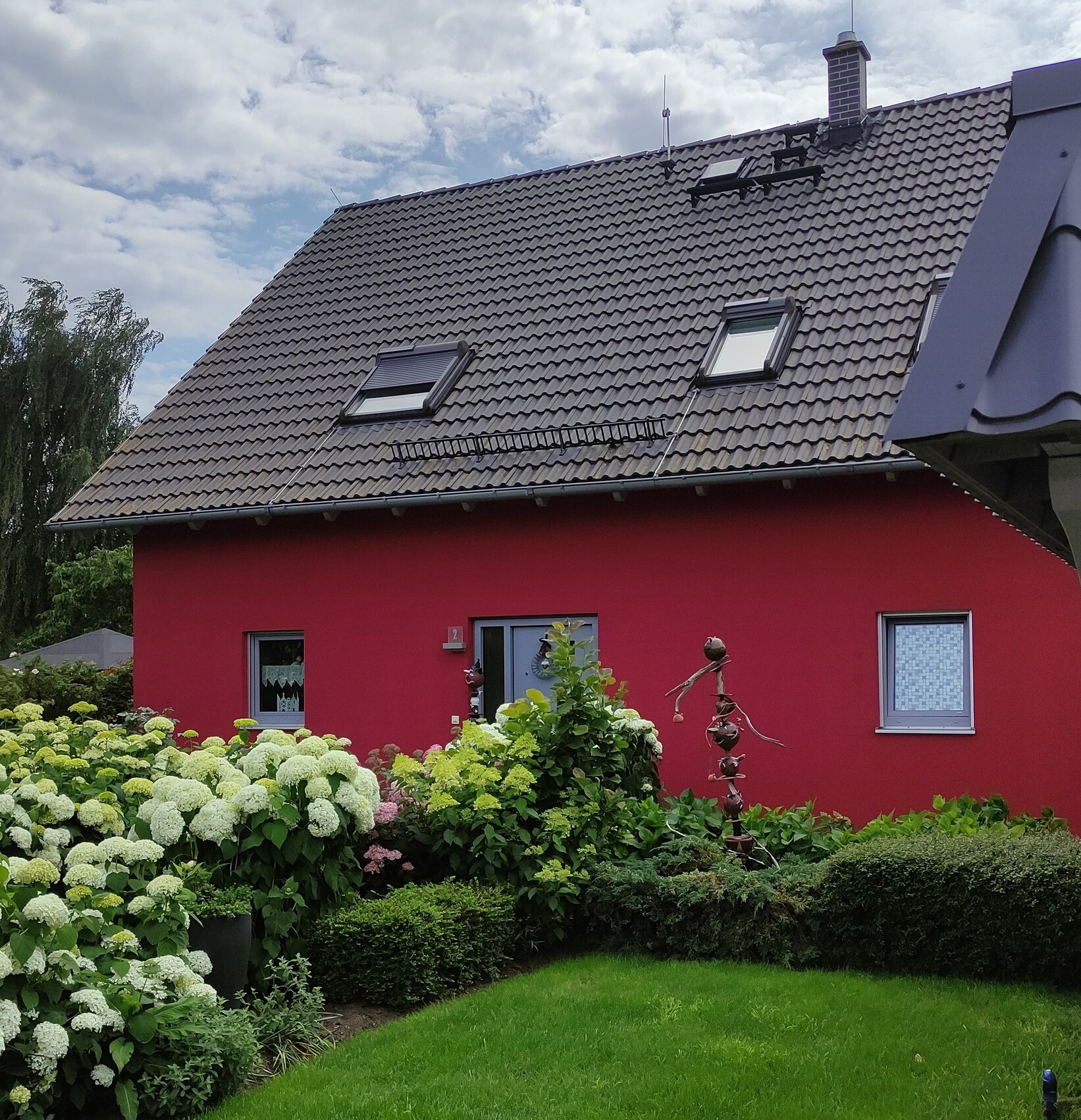 Rotes Haus mit braunem Ziegeldach, zwei Dachfenster, Garten mit Hortensien.