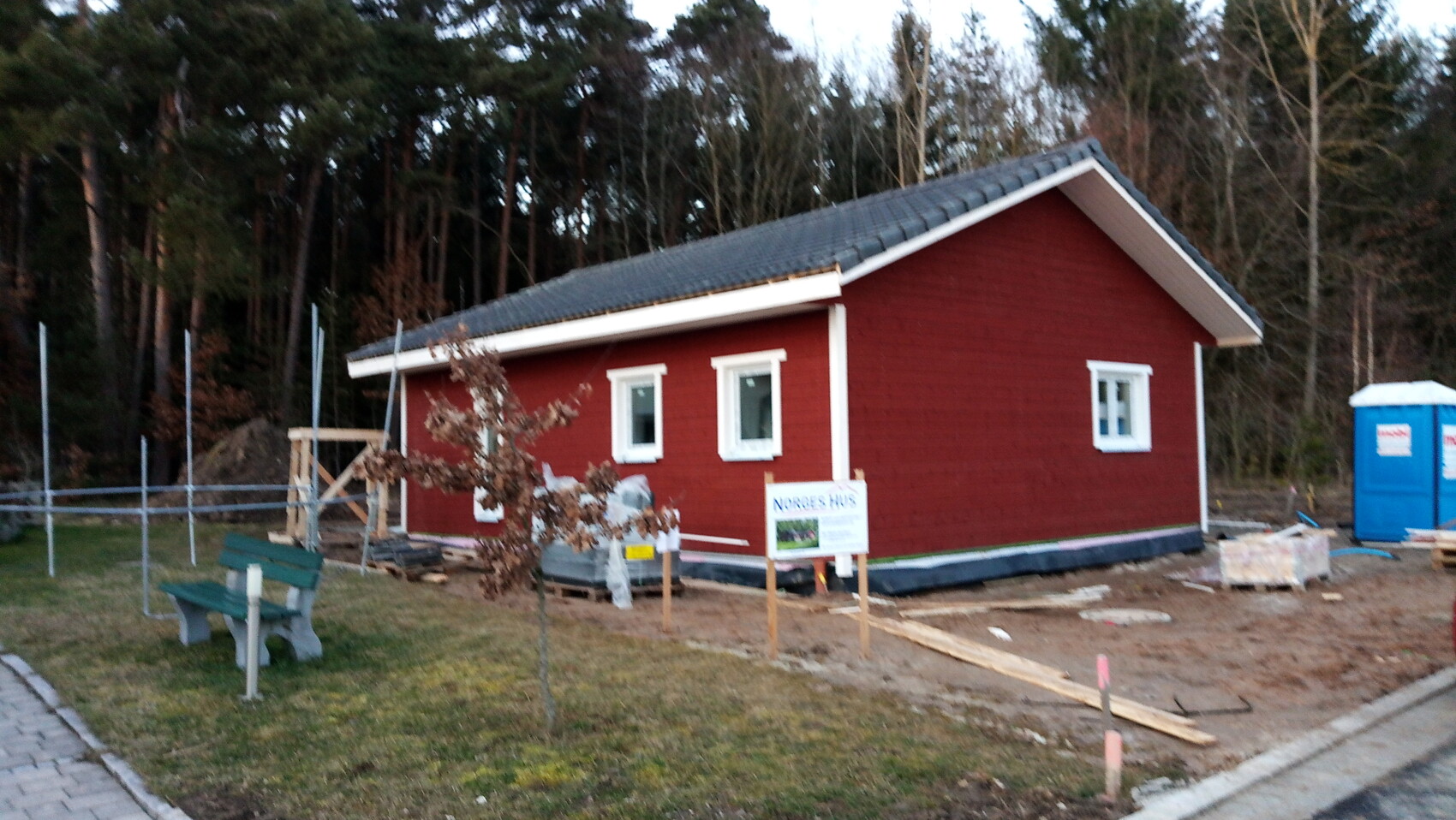 Rotes Holzhaus im Bau am Waldrand, Bauutensilien, Bank und blaue Toilette daneben.