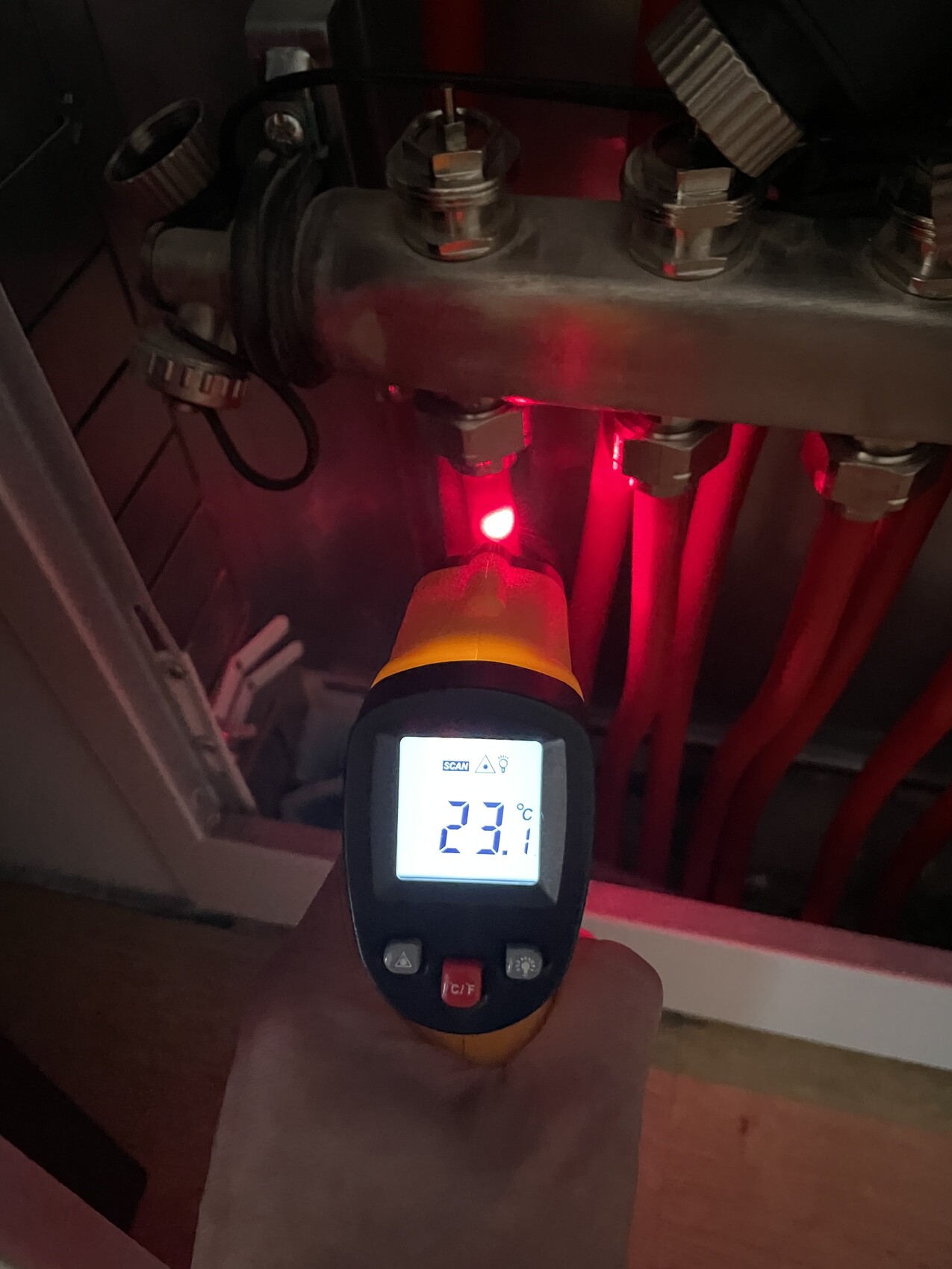 Person benutzt Infrarot-Thermometer, misst Temperatur an rotem Heizungsrohr; Anzeige 23.1 °C.