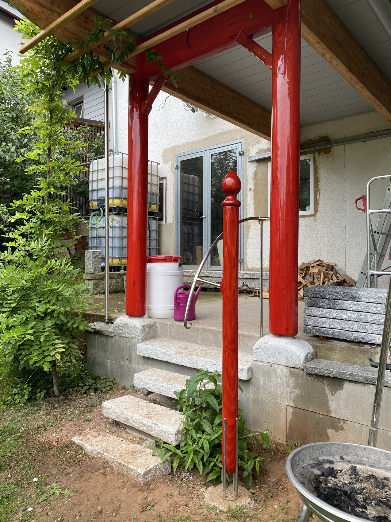 Außenbereich mit roten Holzsaeulen, Treppenstufen, Bauholz und Gartenpflanzen vor dem Haus.