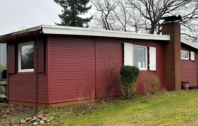 Kleines rotes Holzhaus mit Kamin, weißen Fensterrahmen und Sträuchern davor.