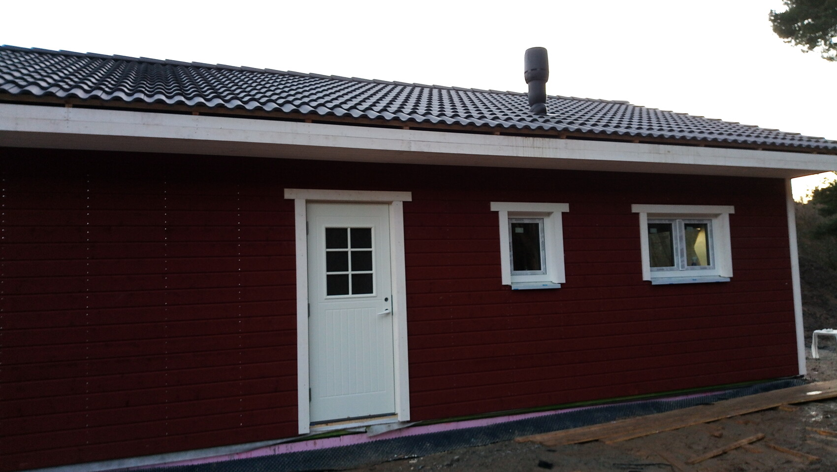 Rotes Holzhaus mit weißer Tür, zwei weißen Fenstern und dunklem Ziegeldach.