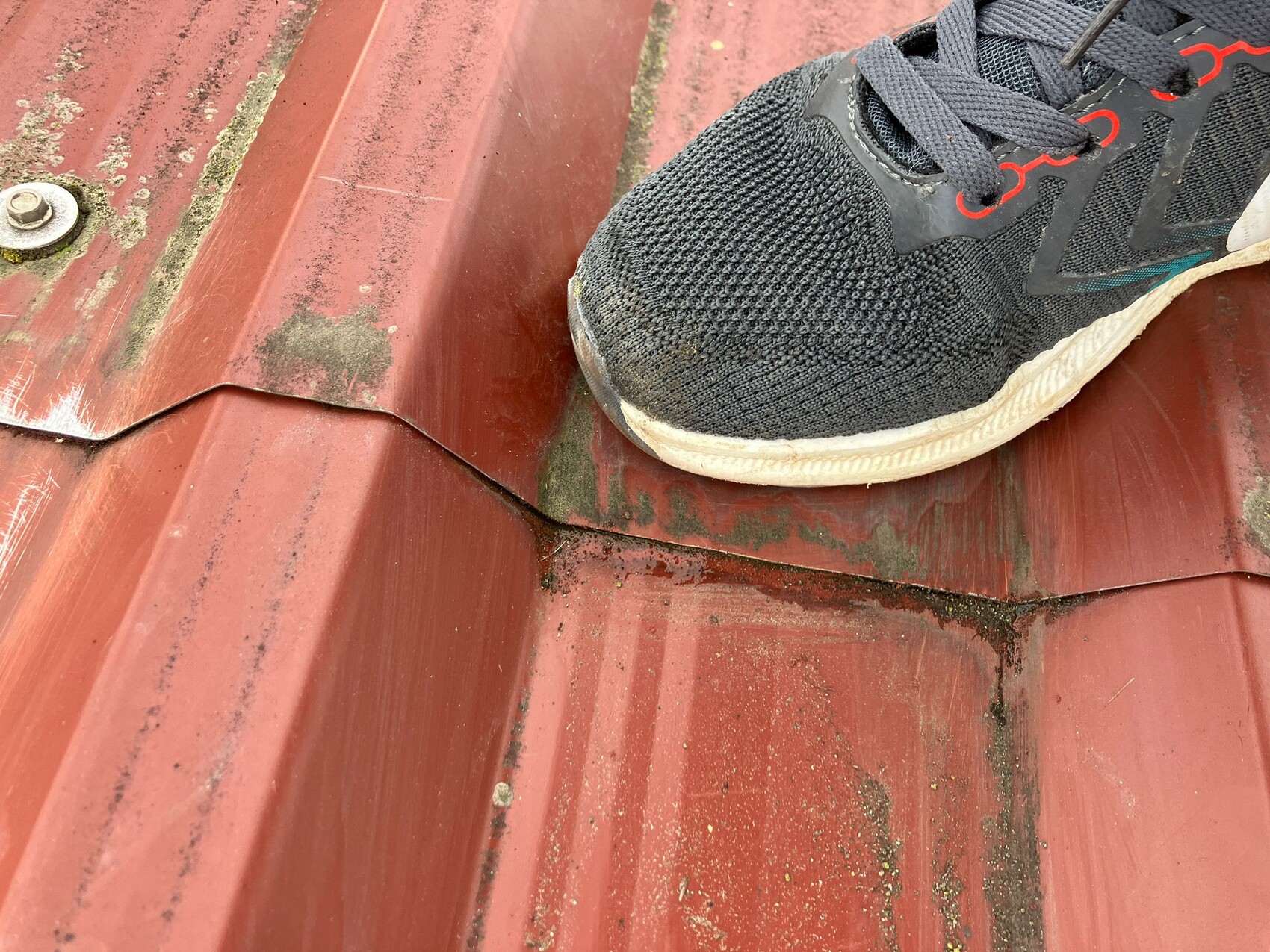 Schwarzer Laufschuh auf rotem Metalldach, Schmutzflecken und Dachnaht sichtbar.