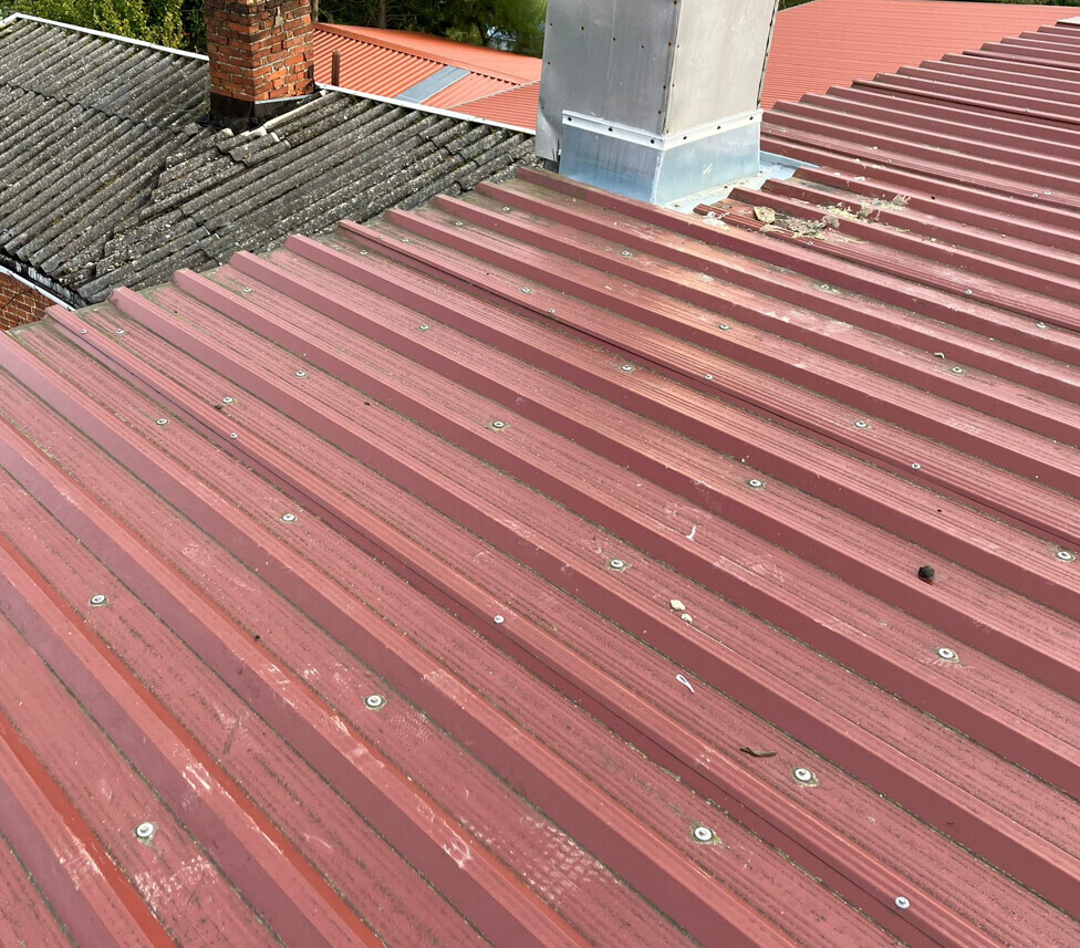 Rotes gewelltes Metalldach mit Schrauben, Schornstein links und Metallaufsatz oben.