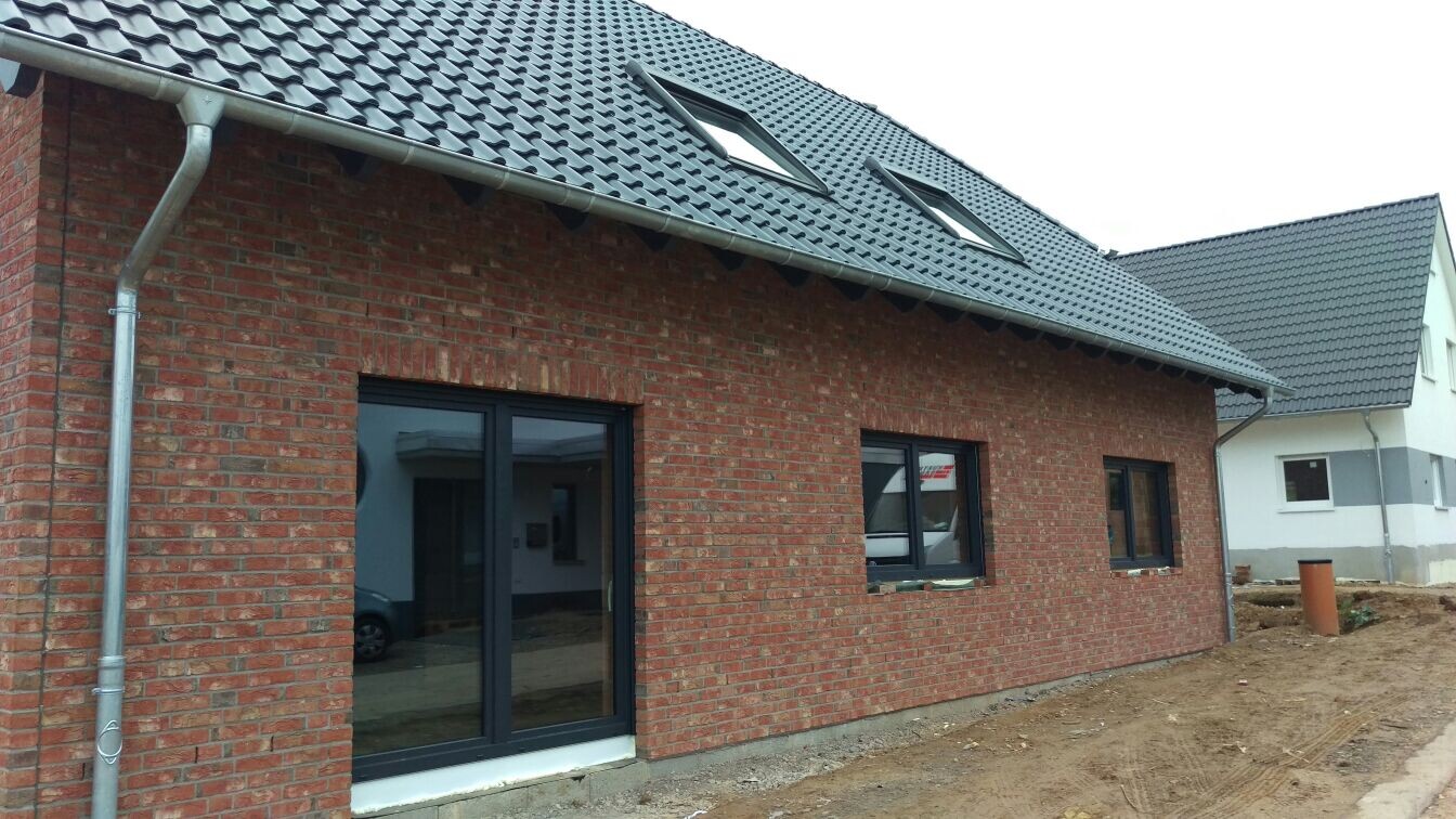Rotes Ziegelhaus im Bau mit großen dunklen Fenstern, Dachfenstern und Baugrund.
