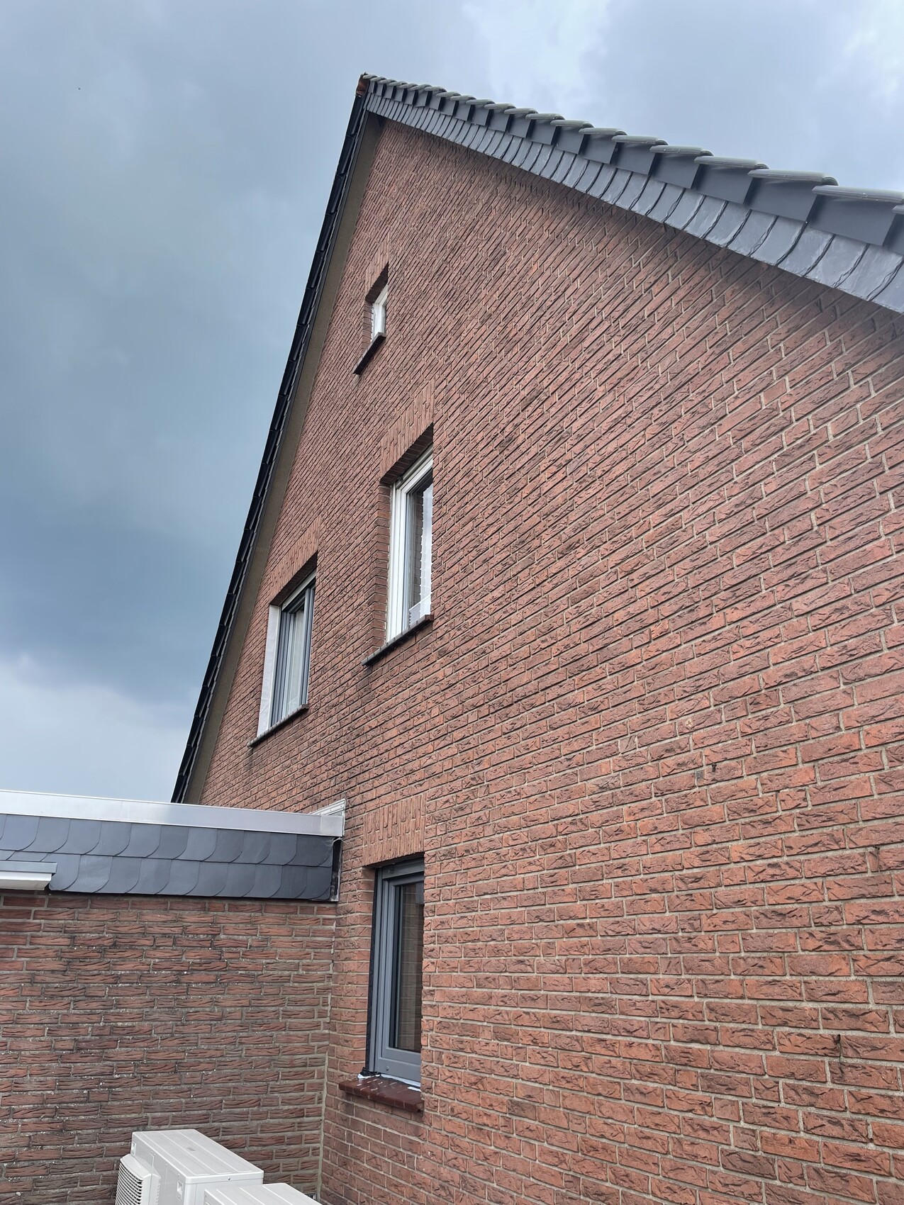 Rotbraunes Ziegelhaus mit schrägem Dach, zwei Fenster und Außenkondensator unten, grauer Himmel