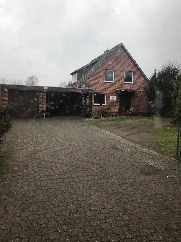 Backsteinhaus mit Satteldach, Carport links, gepflasterter Zufahrtsweg, regnerischer Himmel.