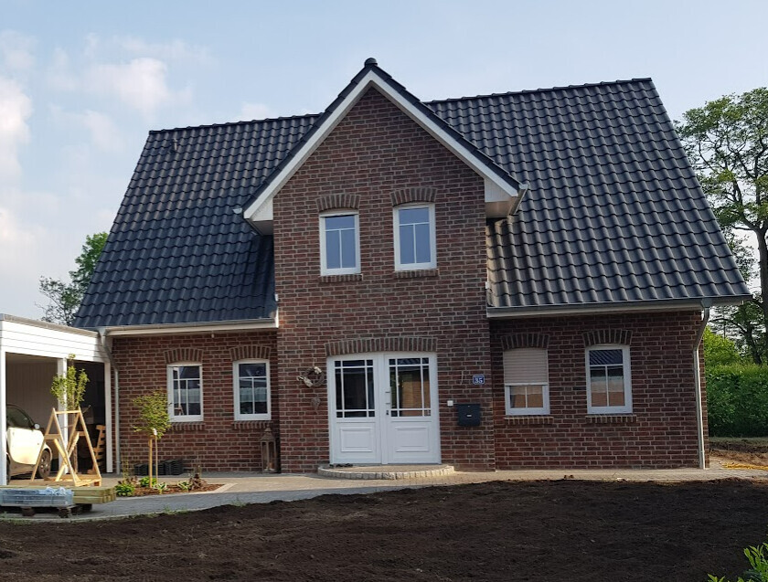 Ziegelhaus mit dunklem Satteldach, weißem Eingang und zwei Fenster darüber, Carport links.