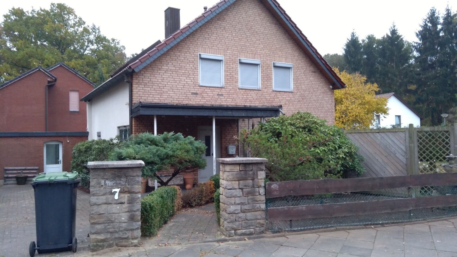 Vorgarten eines zweistöckigen Backsteinhauses mit drei Fenstern, Steinpfeiler Nr. 7, Mülltonne.