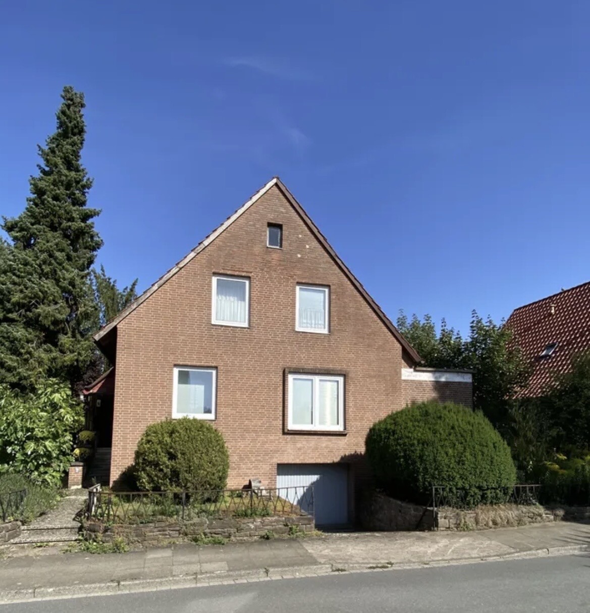 Frontansicht eines roten Ziegelhauses mit Garage, Büschen und grünem Garten vor blauem Himmel.