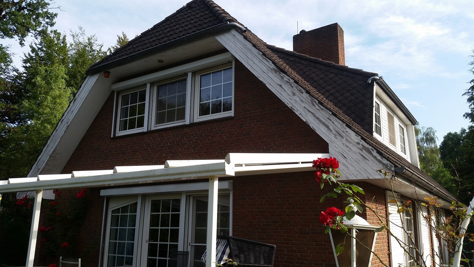 Rotes Backsteinhaus mit dunklem Dach, weiße Pergola und rote Rosen im Garten.