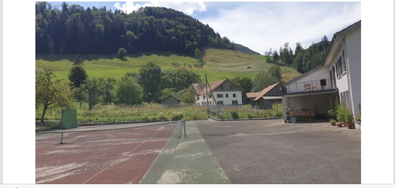 Ländliche Hoflandschaft mit Tennisplatz, Hausreihen und grünem Hang dahinter