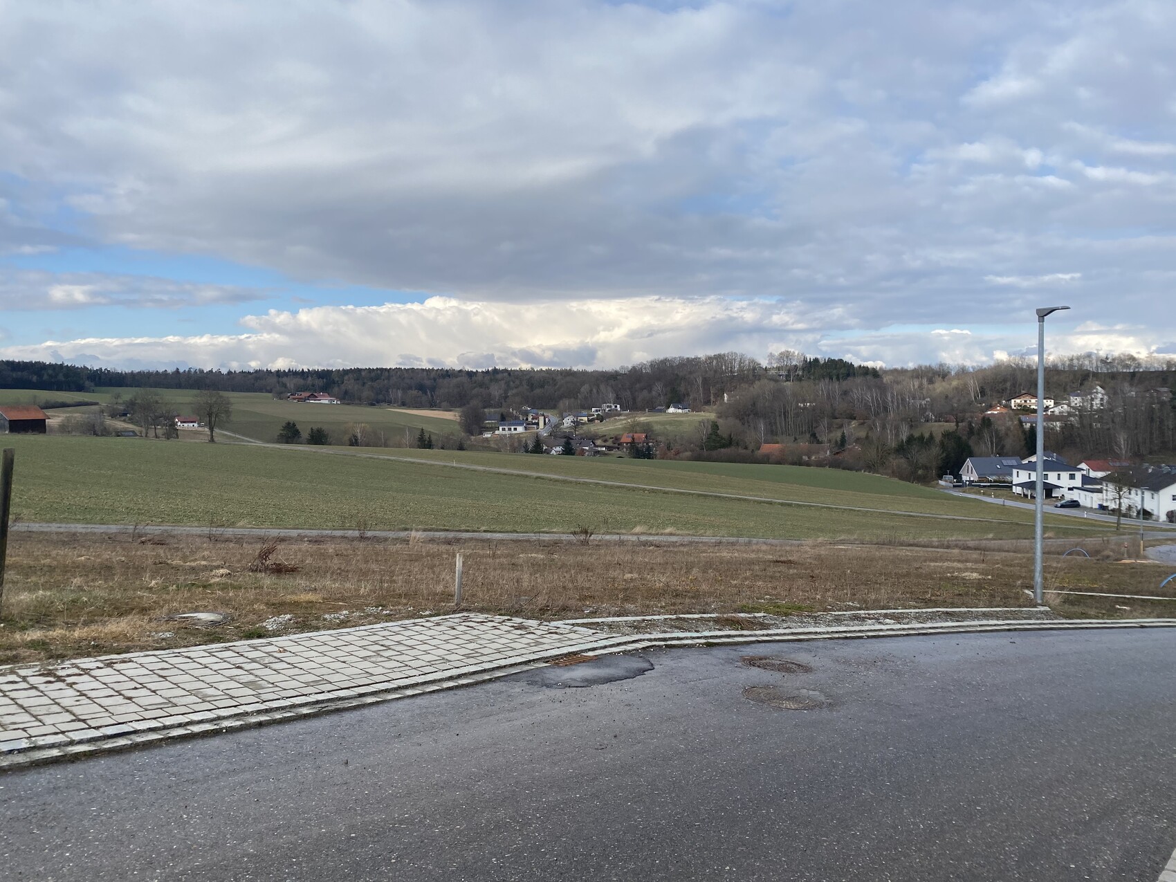 Ländliche Hügellandschaft mit Feldern und Häusern am Hang; Vordergrund: Gehweg und Laterne.