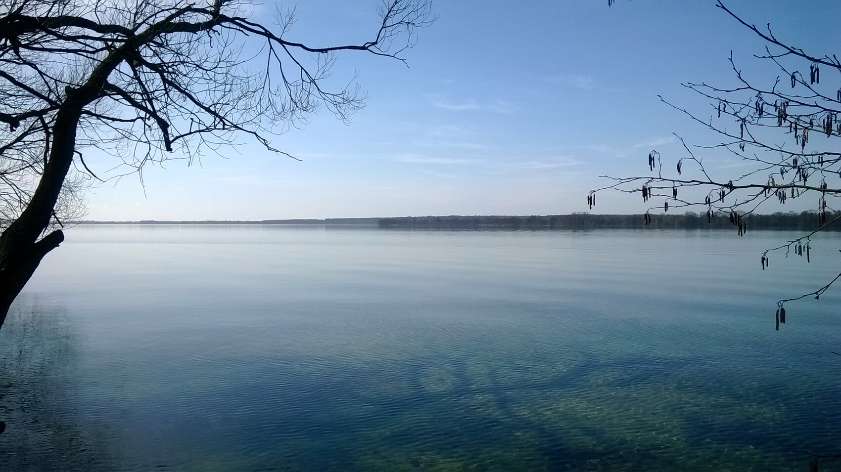 Ruhige Seelandschaft mit klarem Wasser, Uferbäume links und rechts, blauer Himmel.