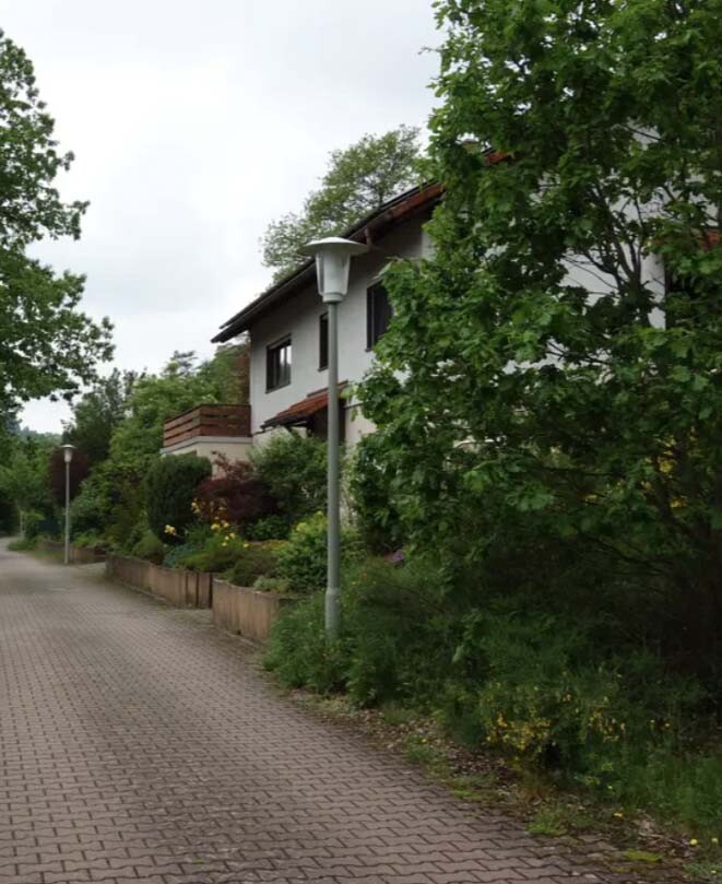Gepflasterter Weg neben einem weißen Wohnhaus, umgeben von Bäumen und einer Straßenlampe.