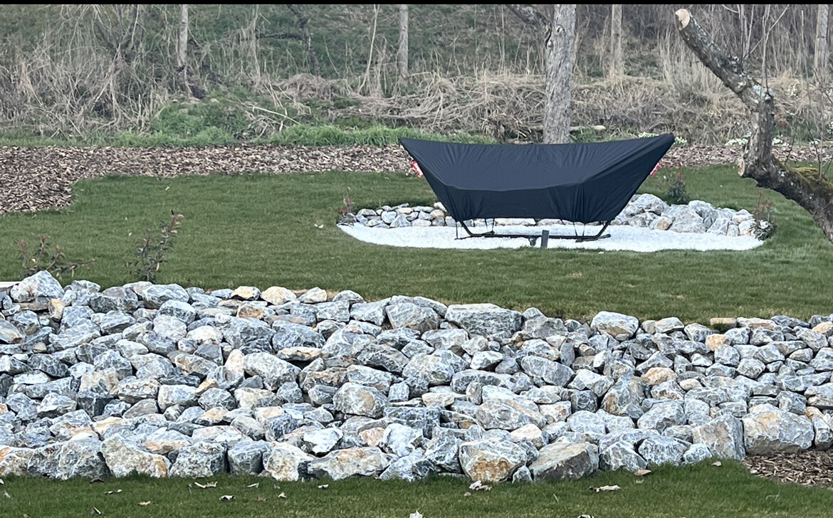 Dunkle Hängematte auf Gestell in einem Kieskreis im Garten, von Gras und Steinmauer umgeben.