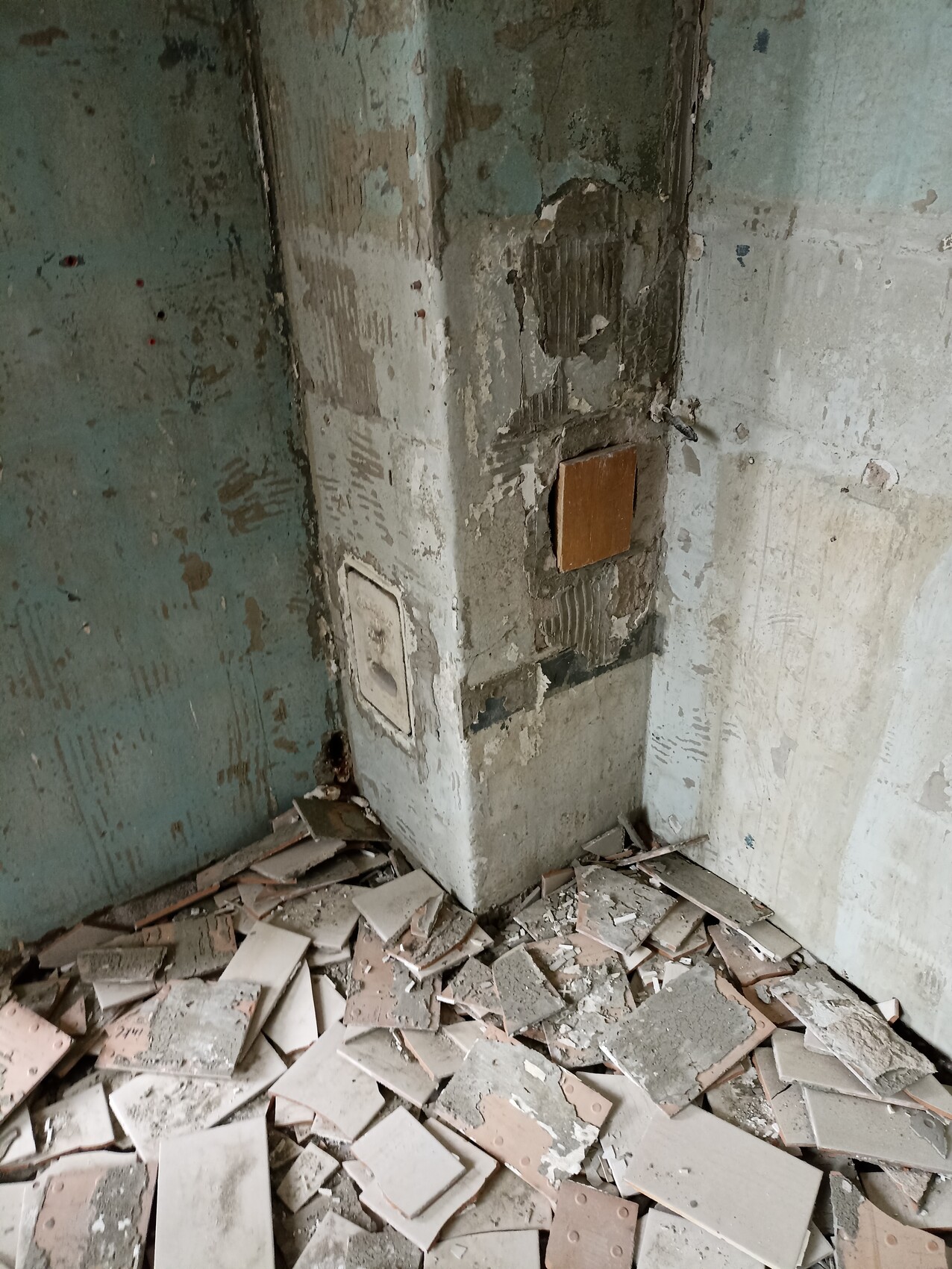 Verlassene Innenraumecke: abblätternde Wand, Betonpfeiler, braune Holzplatte, Fliesenreste auf Boden.
